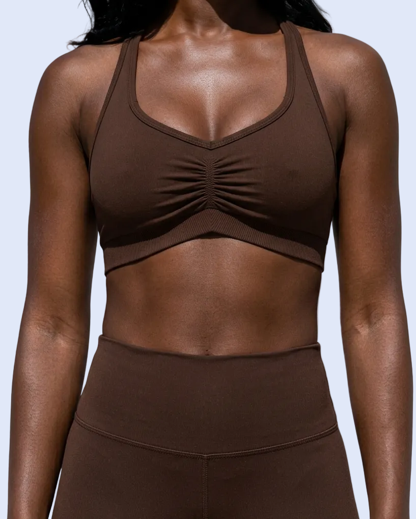A woman wearing a dark brown ruched sports bra and matching high-waisted leggings against a light gray background.