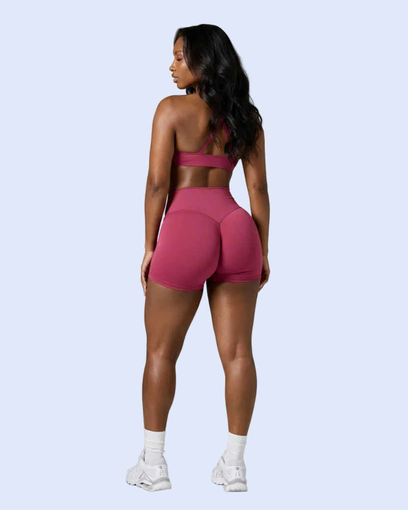 A woman is shown from behind wearing a matching dusty rose pink athletic set, consisting of a sports bra and high-waisted shorts, paired with white socks and sneakers, against a solid gray studio background.