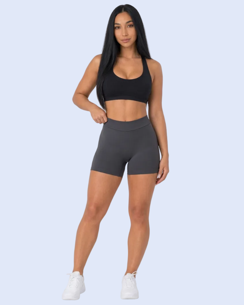 A woman modeling a matching activewear set consisting of a black sports bra and dark gray high-waisted biker shorts, paired with white sneakers, against a light gray studio background.