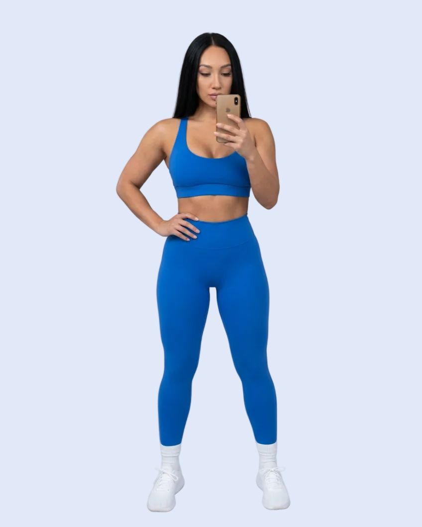 A woman posing in a matching royal blue activewear set consisting of a sports bra and high-waisted leggings, paired with white socks and sneakers, holding a gold smartphone in front of her face against a light gray studio background.
