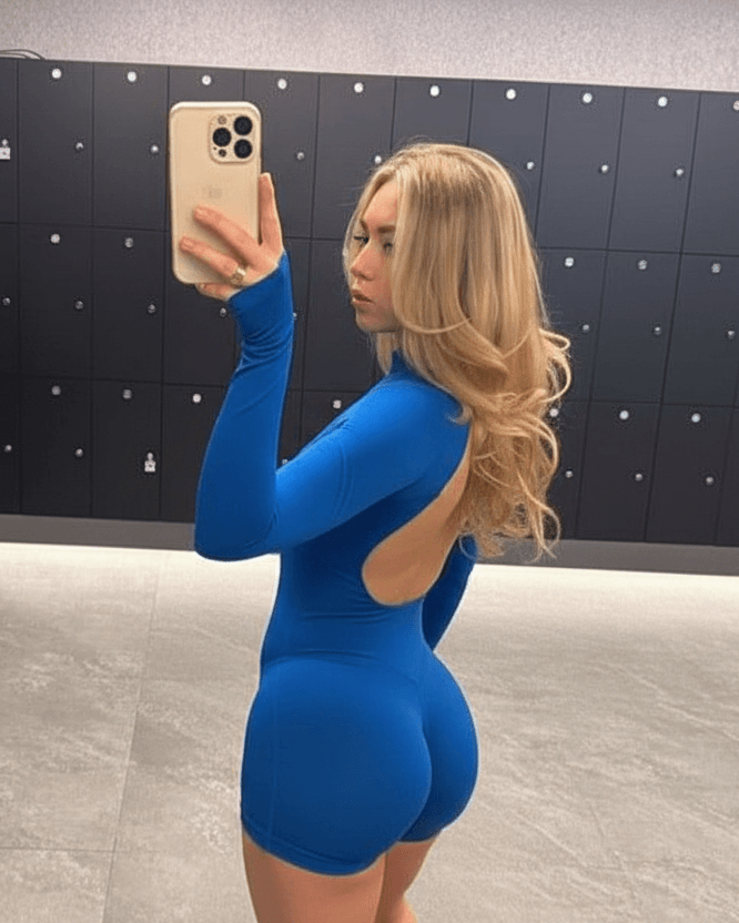 A blonde woman takes a mirror selfie in a locker room wearing a bright blue long-sleeve mini dress with an open back. She is holding a gold-colored smartphone, with dark gray lockers and a light gray tiled floor in the background.