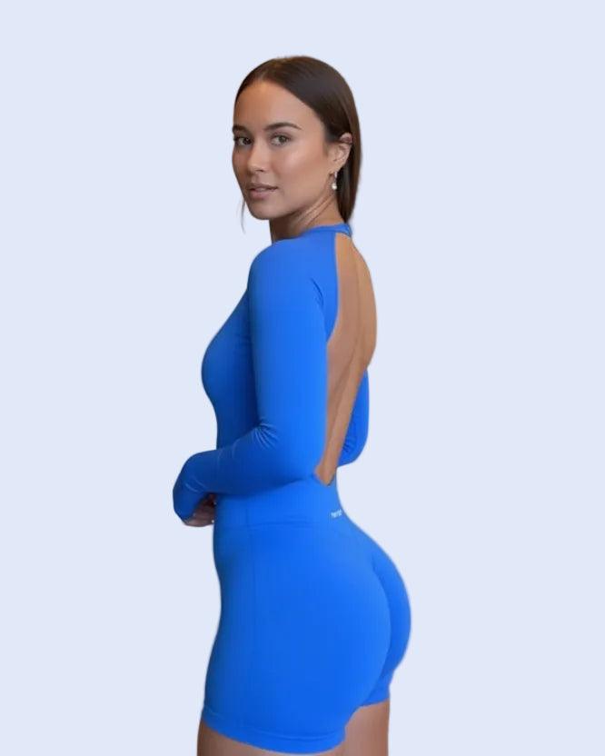 A woman wearing a bright blue long-sleeve mini bodycon dress with a large open back, posing in a modern kitchen interior.