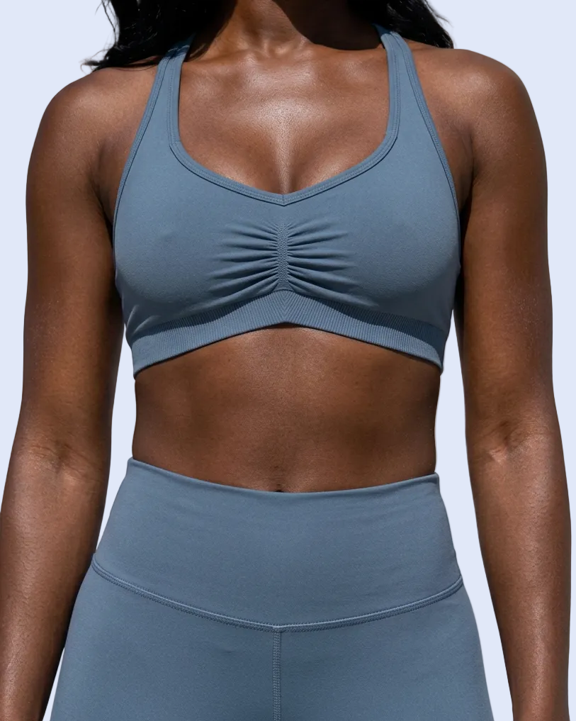 A woman wearing a slate blue-gray sports bra with a ruched center detail and matching high-waisted leggings against a light gray background.