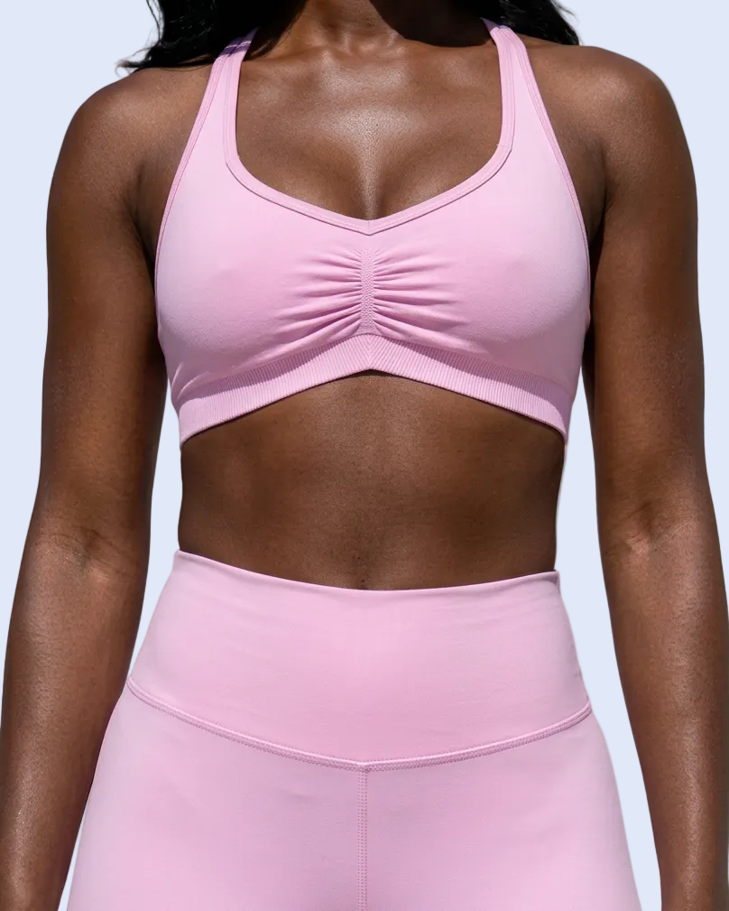 A woman wearing a light pink athletic sports bra with a ruched center detail and matching high-waisted leggings, shown from the chest to the hips against a light gray studio background.