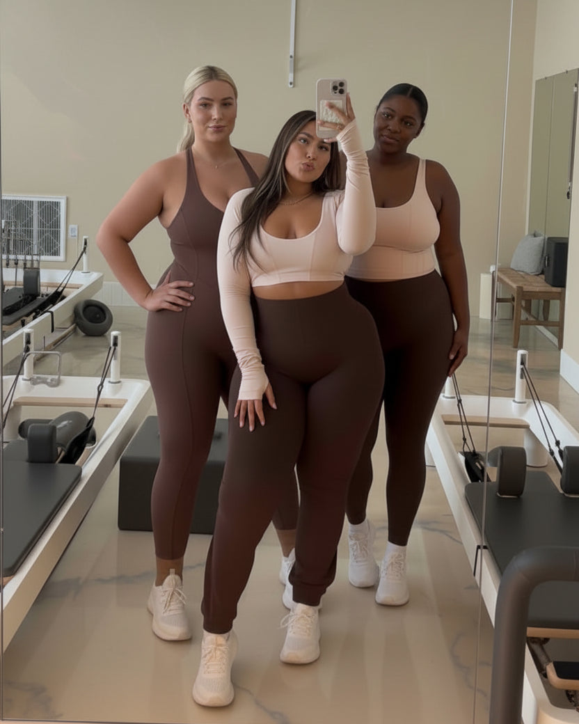 Three women posing together in a fitness studio wearing matching athletic wear in shades of taupe, beige, and dark brown. The woman in the center is taking a mirror selfie with a phone.