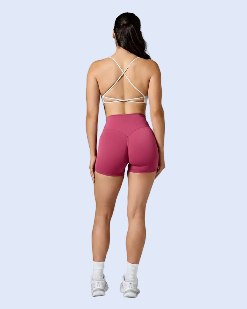 A woman is shown from behind wearing a white strappy sports bra and high-waisted magenta athletic shorts, standing against a neutral gray studio background.