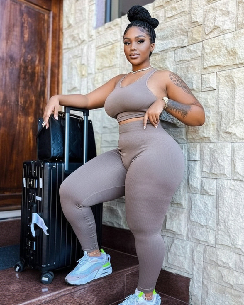 A woman posing in a matching ribbed activewear set consisting of a sports bra and high-waisted leggings in taupe, standing next to a black suitcase against a stone wall.