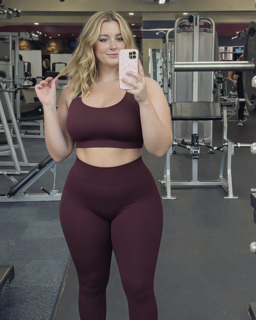 A woman takes a mirror selfie in a gym while wearing a matching burgundy sports bra and high-waisted leggings set.