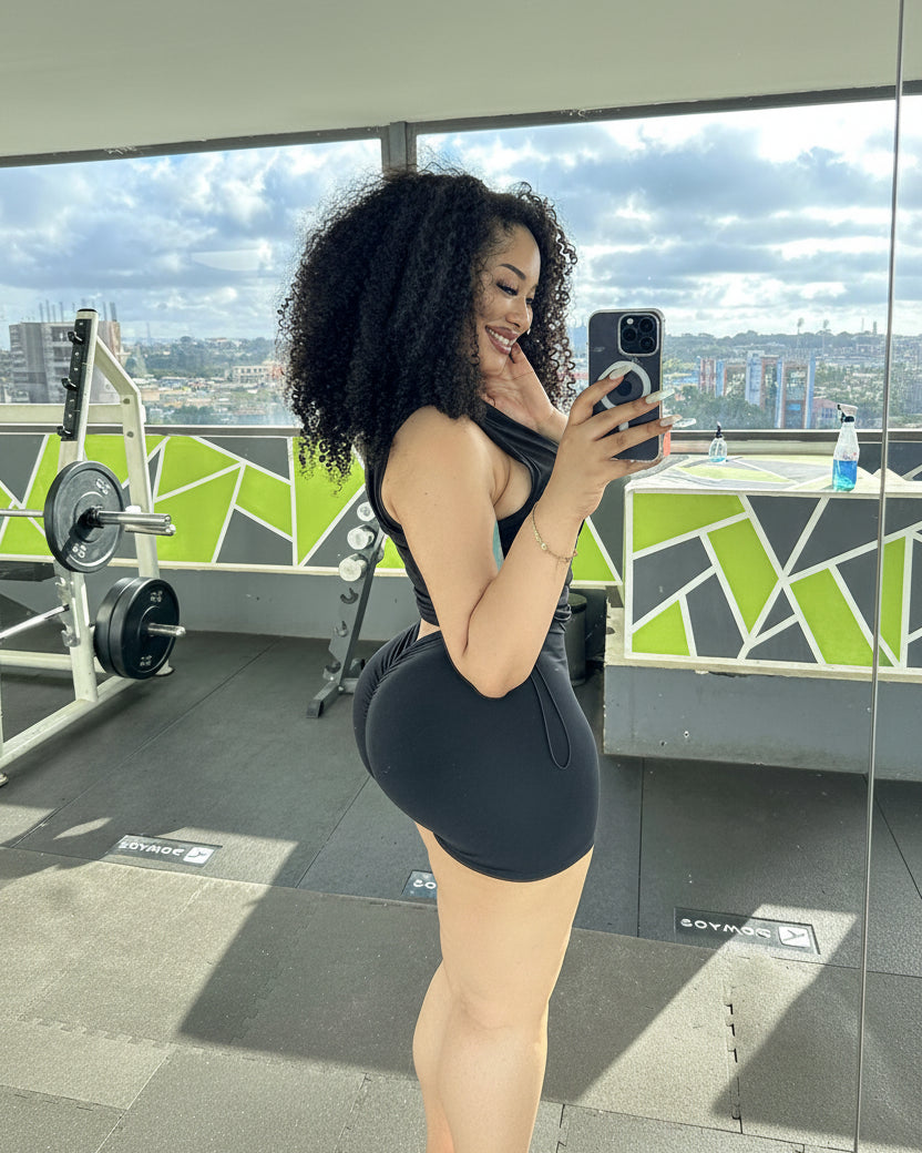 A woman takes a mirror selfie in a gym while wearing a black athletic bodysuit. The background shows gym equipment, a green and gray geometric wall, and a city skyline visible through large windows.