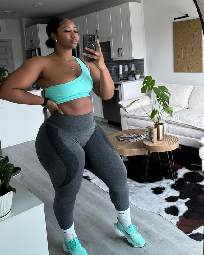 A woman takes a mirror selfie in a modern living room, wearing a two-piece activewear set consisting of a light turquoise one-shoulder crop top and matching high-waisted gray leggings.
