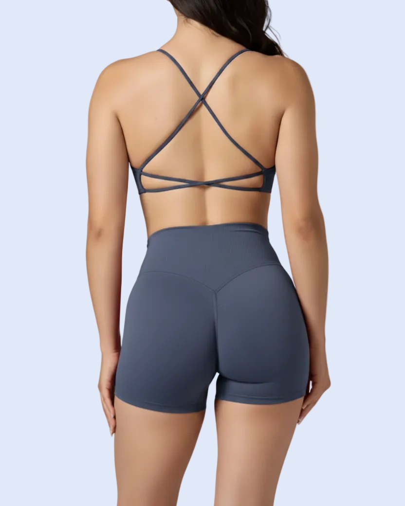 Rear view of a woman wearing a dark slate blue sports bra with crisscross back straps and matching high-waisted athletic shorts against a light blue background.