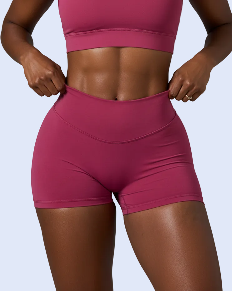 A model wearing a matching athletic set consisting of a scoop-neck sports bra and high-waisted shorts in a dusty rose or magenta color, shown against a solid gray studio background.