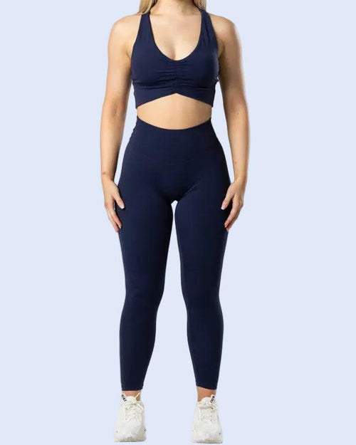 A woman modeling a matching navy blue activewear set consisting of a sports bra and high-waisted leggings, paired with white sneakers, against a light gray studio background.