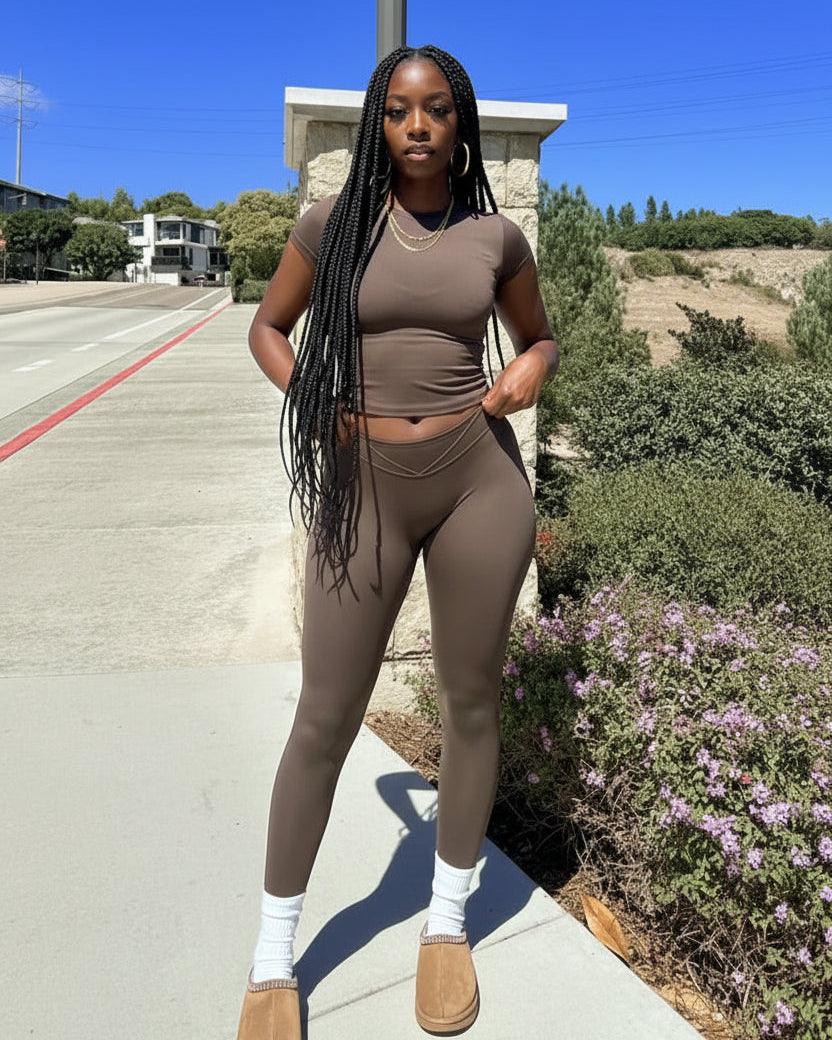 A woman stands outdoors on a sidewalk wearing a fitted short-sleeve brown mock-neck top and matching high-waisted brown leggings, with white socks and tan slip-on shoes. The background shows a road, stone wall, shrubs, and blue sky.